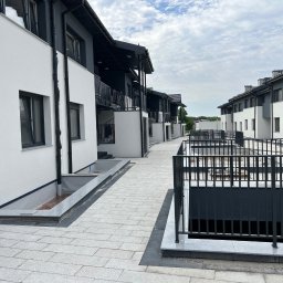 ITB Biuro Inżynierskie Piotr Kurasiński - Nowoczesne szeregowe domy z balkonami, białe elewacje z szarymi akcentami, chodnik wyłożony jasną kostką brukową, barierki balkonowe z czarnych prętów.