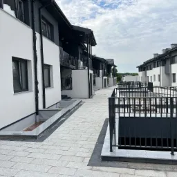 Nowoczesne szeregowe domy z balkonami, białe elewacje z szarymi akcentami, chodnik wyłożony jasną kostką brukową, barierki balkonowe z czarnych prętów.