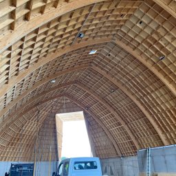 ITB Biuro Inżynierskie Piotr Kurasiński - Konstrukcja drewniana dachu hali w trakcie budowy, widoczne elementy kratownicy i deskowanie, w tle częściowo widoczny samochód dostawczy.