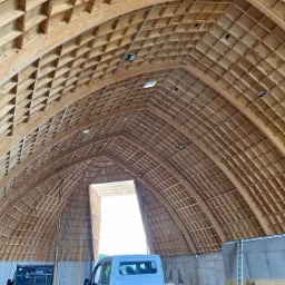 Konstrukcja drewniana dachu hali w trakcie budowy, widoczne elementy kratownicy i deskowanie, w tle częściowo widoczny samochód dostawczy.