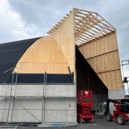 ITB Biuro Inżynierskie Piotr Kurasiński - Budowa hali z elementami drewnianymi i betonowymi ścianami, widoczne rusztowania, podnośniki Manitou i pracownik w odblaskowej kamizelce, dach częściowo pokryty czarną papą.