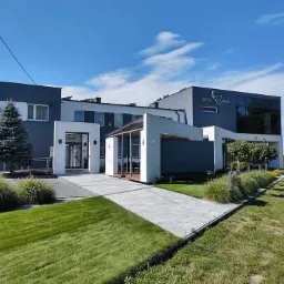 Nowoczesny budynek hotelu z szaro-białą elewacją, widok z zewnątrz w słoneczny dzień, zadbany trawnik i krzewy ozdobne.