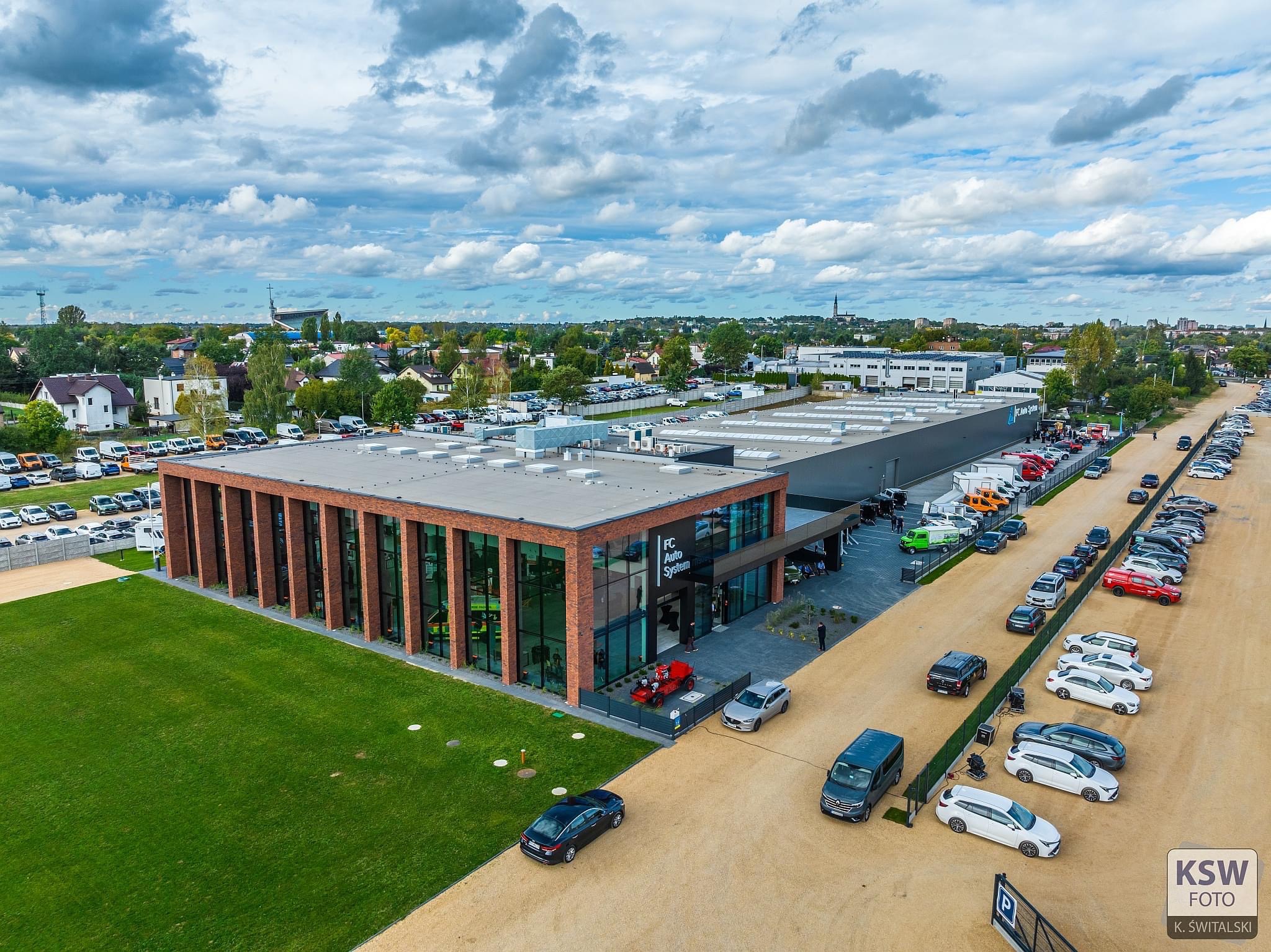 Zdjęcie z lotu ptaka nowoczesnego budynku biurowo-usługowego z elementami cegły, z dużym parkingiem i zadbanym trawnikiem.