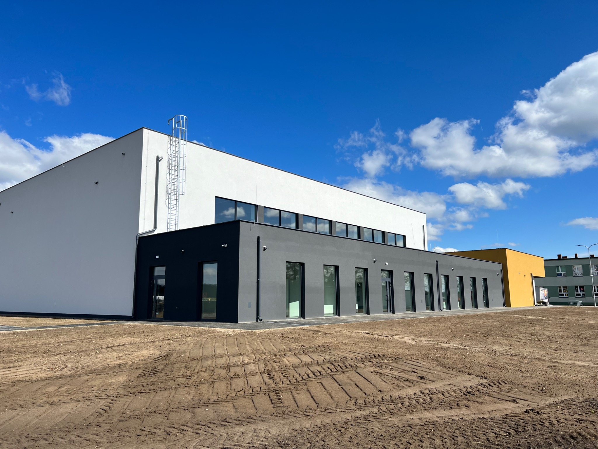 Nowoczesny budynek przemysłowy z białymi i szarymi ścianami, z zewnętrzną drabiną na dachu, na tle błękitnego nieba z chmurami, teren wokół budynku przygotowany pod zagospodarowanie.