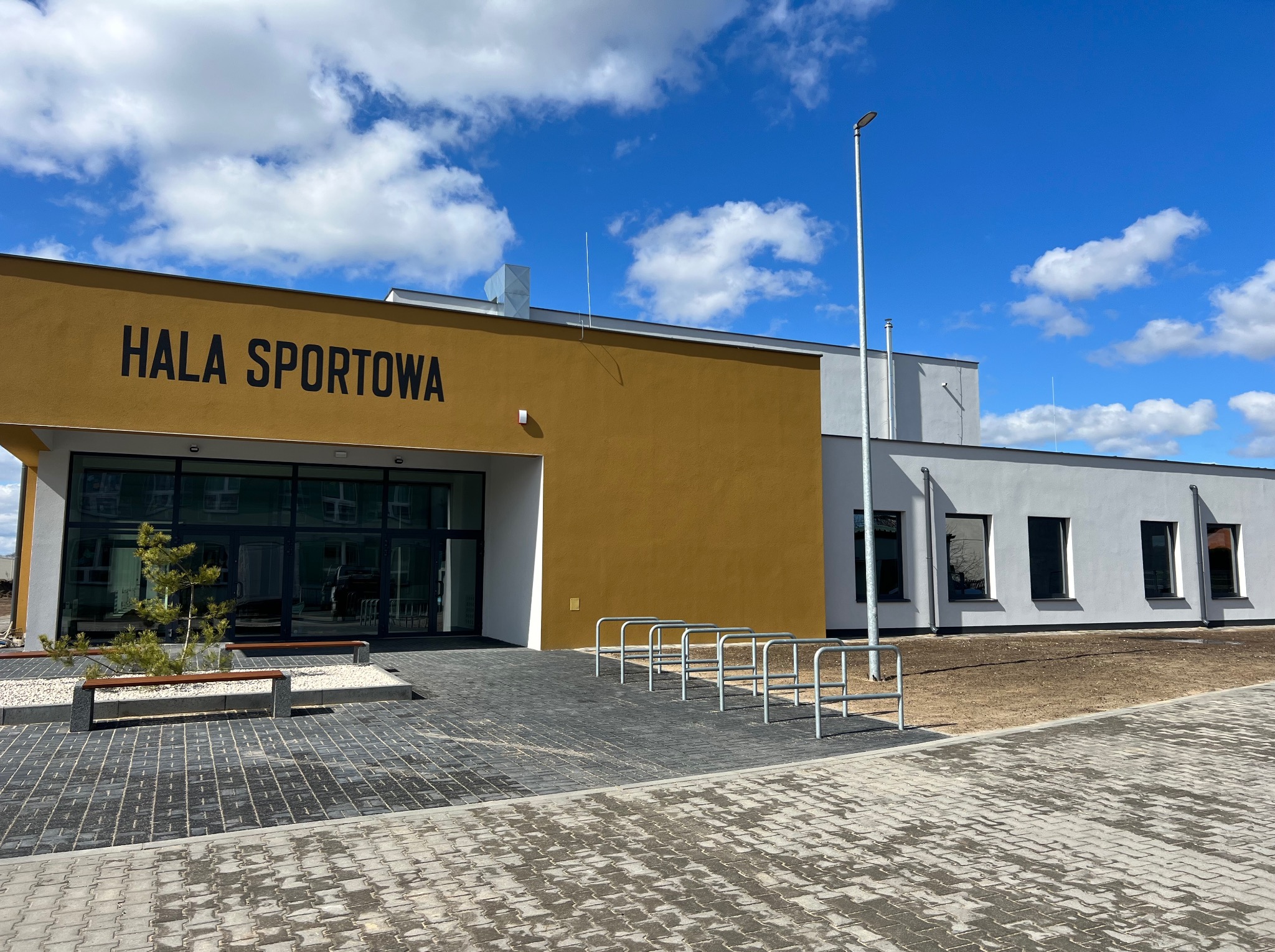 Nowoczesna hala sportowa z żółtą elewacją wejściową i napisem 'HALA SPORTOWA', widoczny chodnik z kostki brukowej, ławki i stojaki na rowery, błękitne niebo z chmurami w tle.