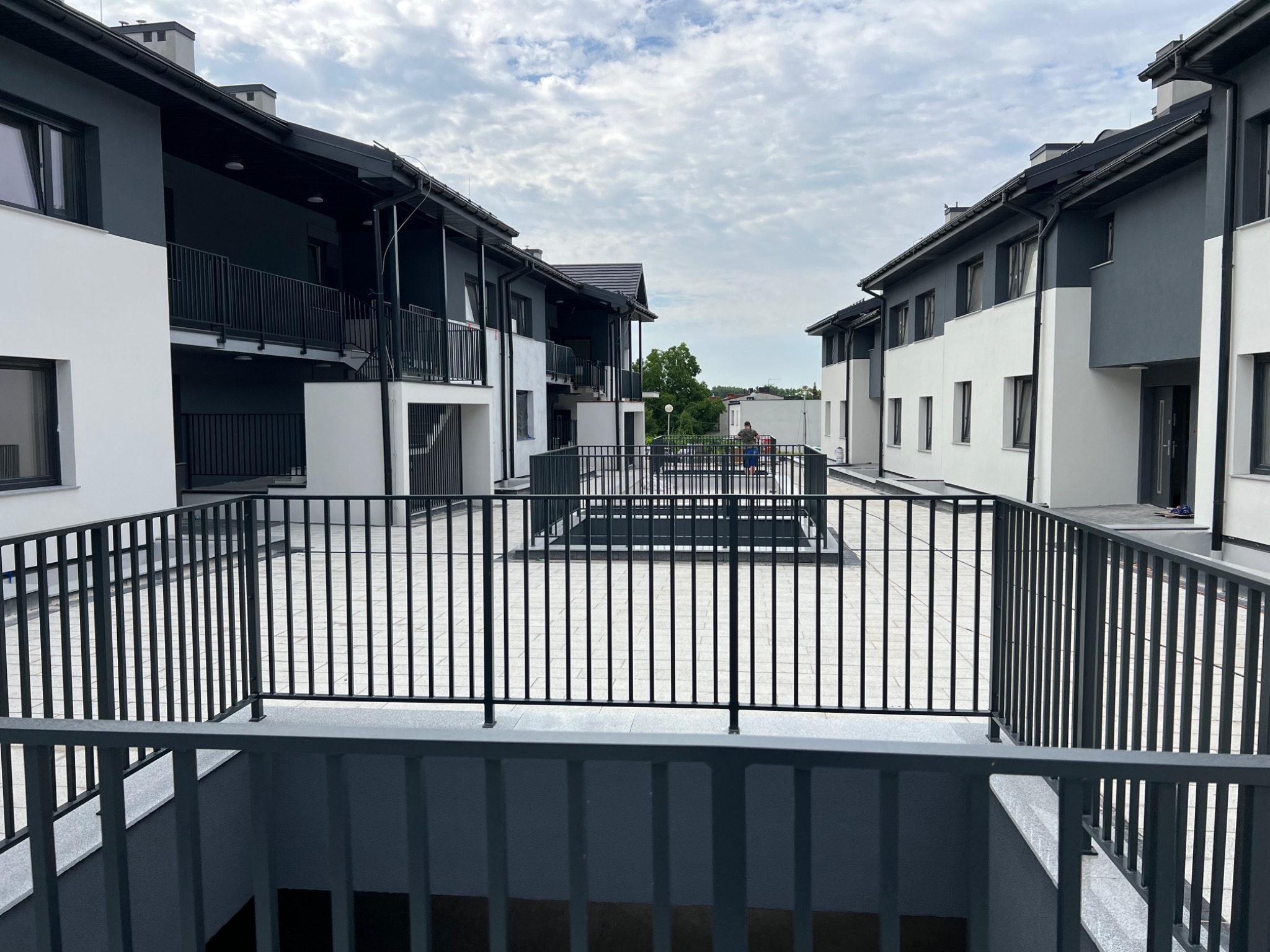 Nowoczesne osiedle budynków wielorodzinnych z balkonami i wewnętrznym dziedzińcem wyłożonym kostką, widok z wyższego poziomu na metalowe balustrady i osobę siedzącą w oddali.