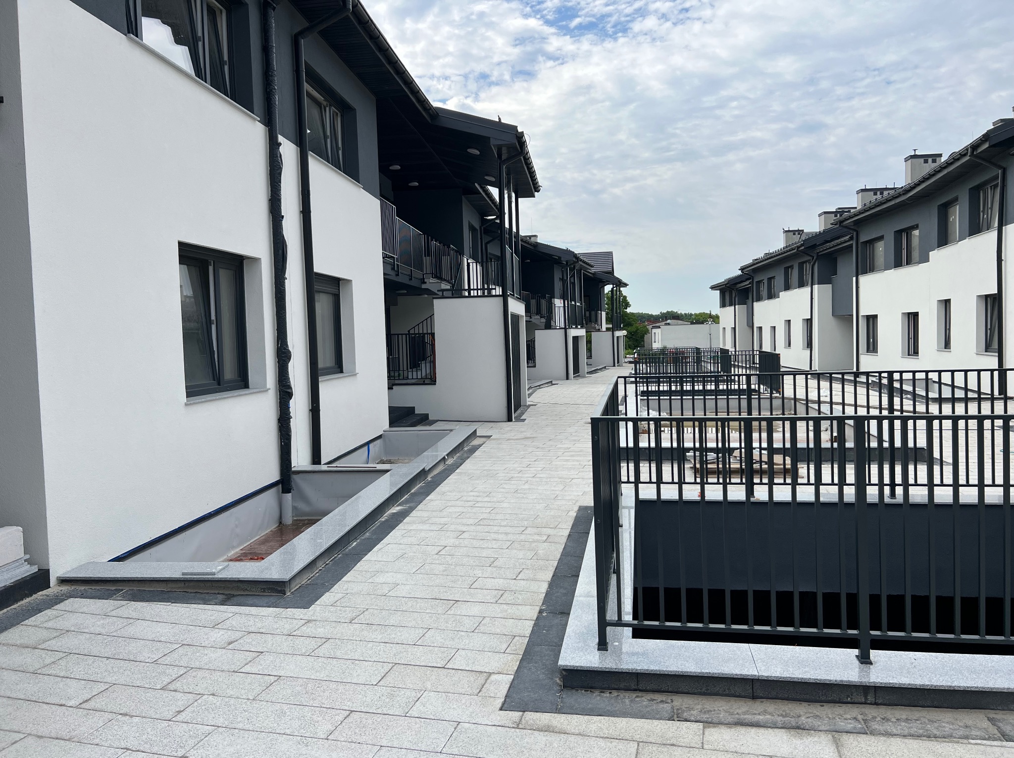 Nowoczesne szeregowe domy z balkonami, białe elewacje z szarymi akcentami, chodnik wyłożony jasną kostką brukową, barierki balkonowe z czarnych prętów.