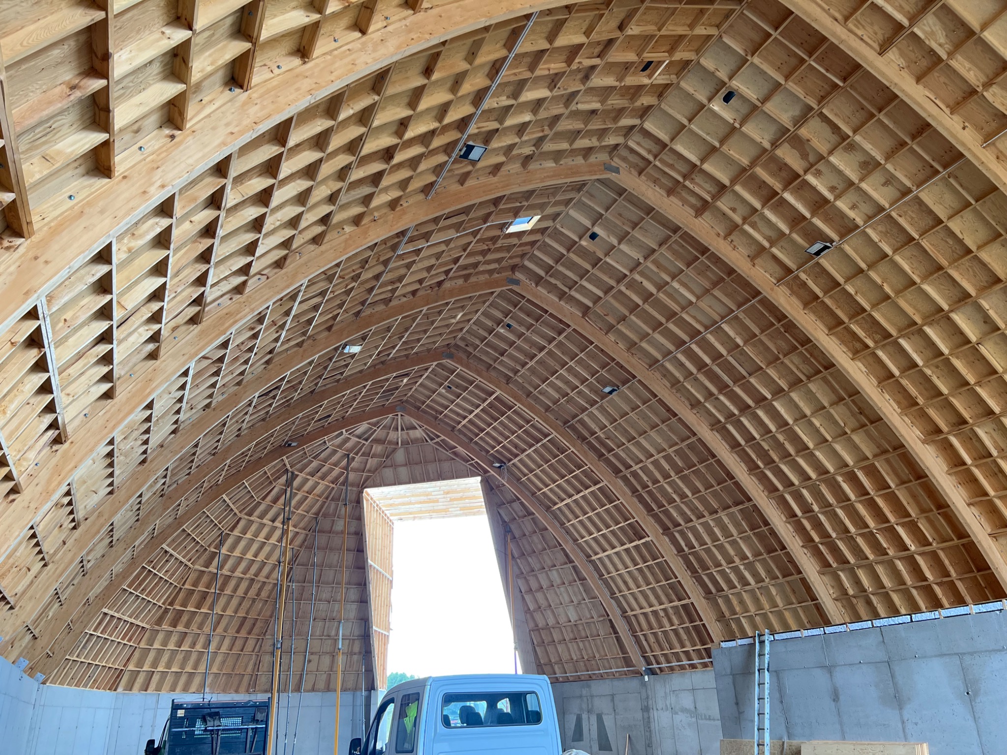 Konstrukcja drewniana dachu hali w trakcie budowy, widoczne elementy kratownicy i deskowanie, w tle częściowo widoczny samochód dostawczy.