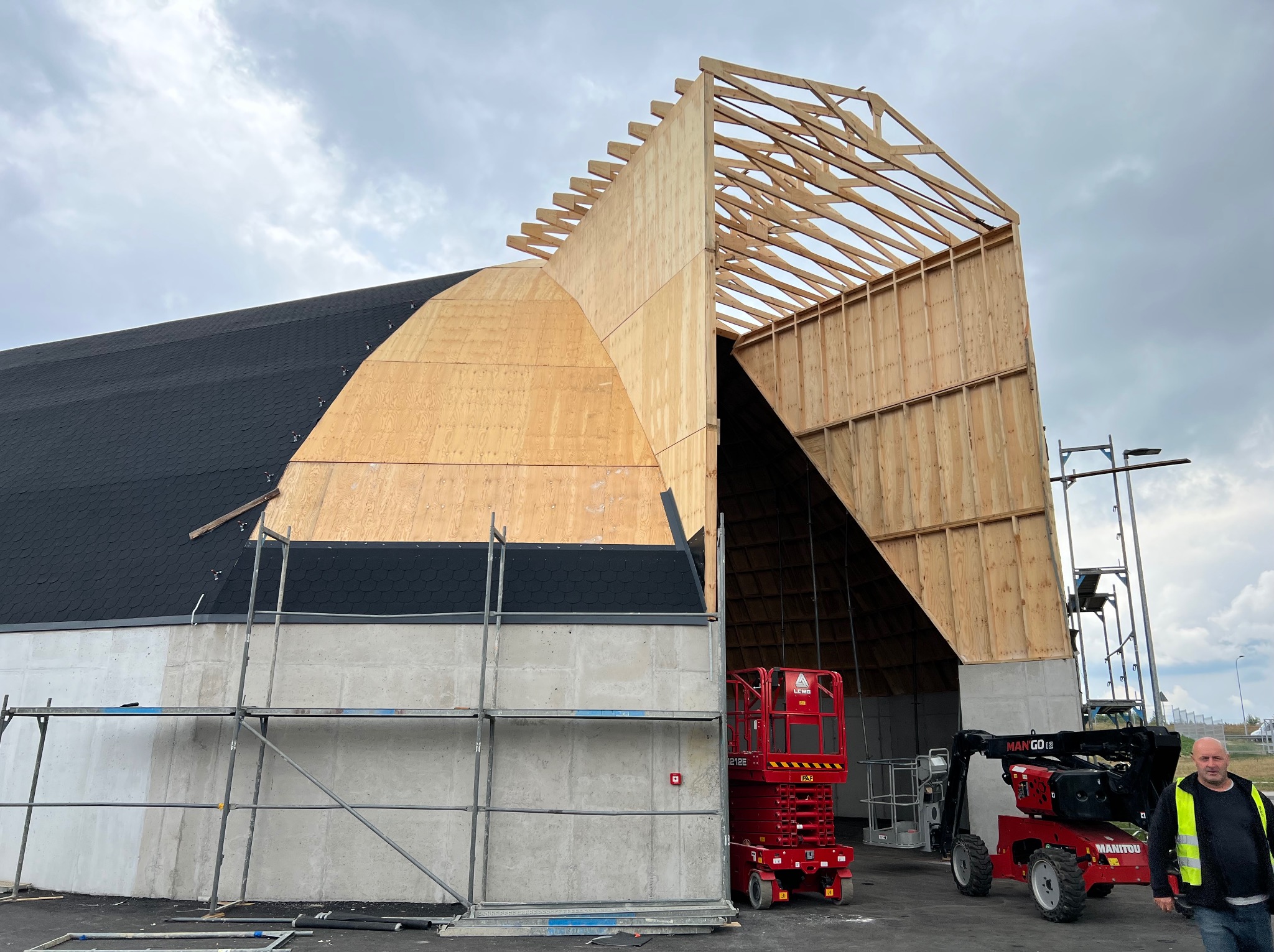 Budowa hali z elementami drewnianymi i betonowymi ścianami, widoczne rusztowania, podnośniki Manitou i pracownik w odblaskowej kamizelce, dach częściowo pokryty czarną papą.