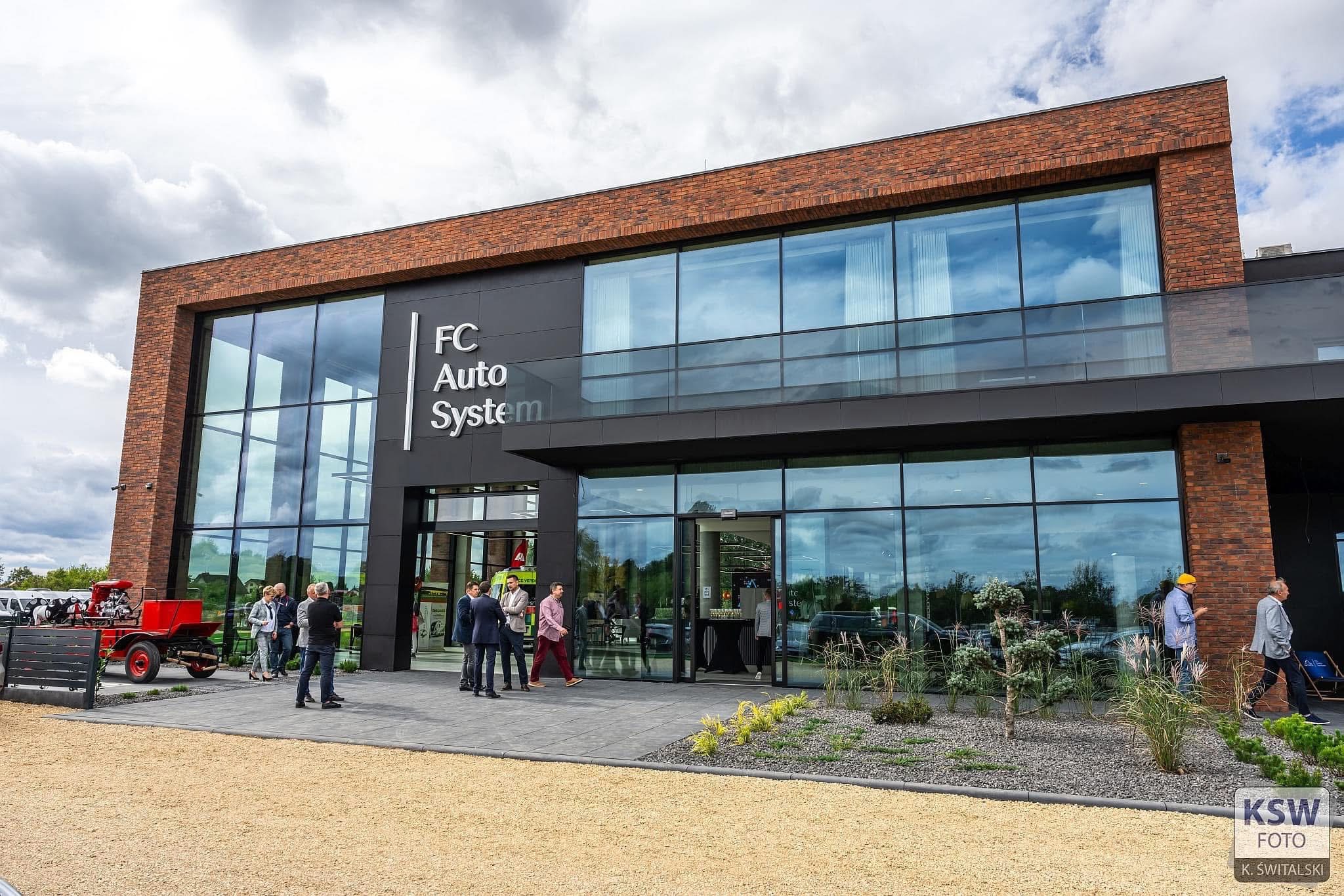 Nowoczesny budynek biurowy z dużymi oknami i ceglanym wykończeniem, grupa ludzi przed wejściem, czerwony wóz strażacki jako element dekoracyjny.