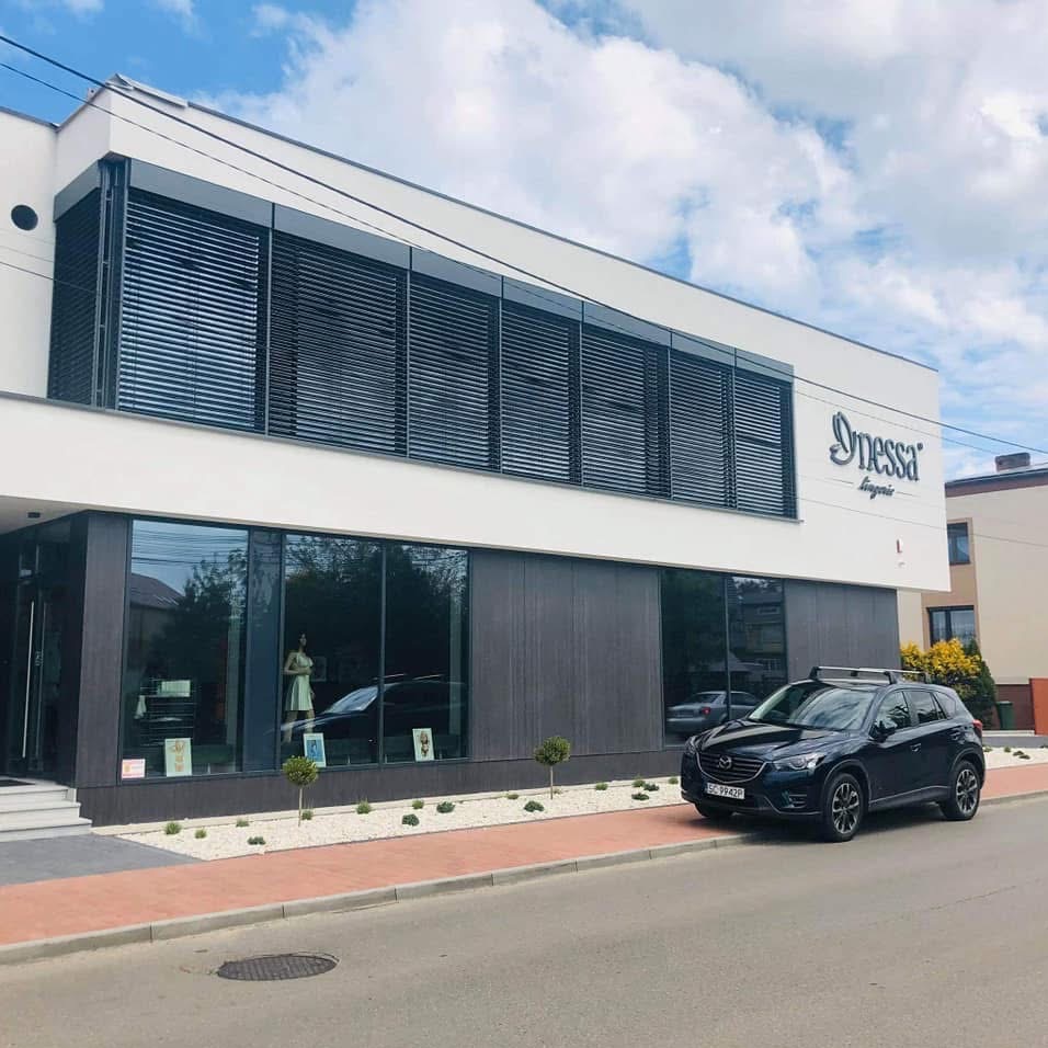 Nowoczesny budynek usługowy z dużymi oknami, żaluzjami zewnętrznymi i zaparkowanym samochodem osobowym przed wejściem.