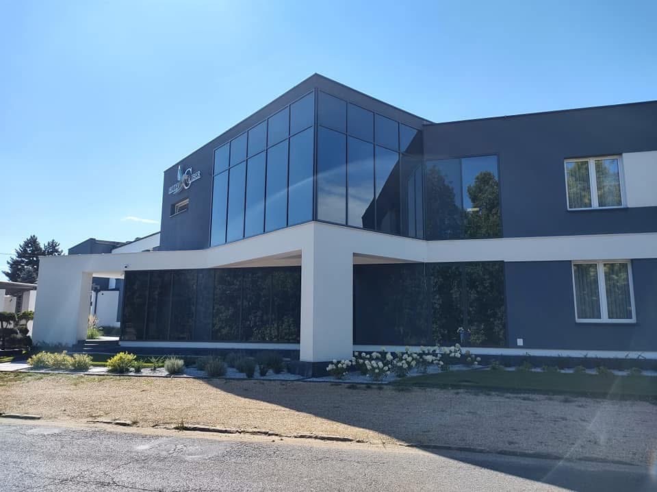 Nowoczesny budynek usługowy z dużymi przeszkleniami i białymi elementami architektonicznymi, otoczony zielenią i kwiatami. Widoczny znak firmowy na elewacji.