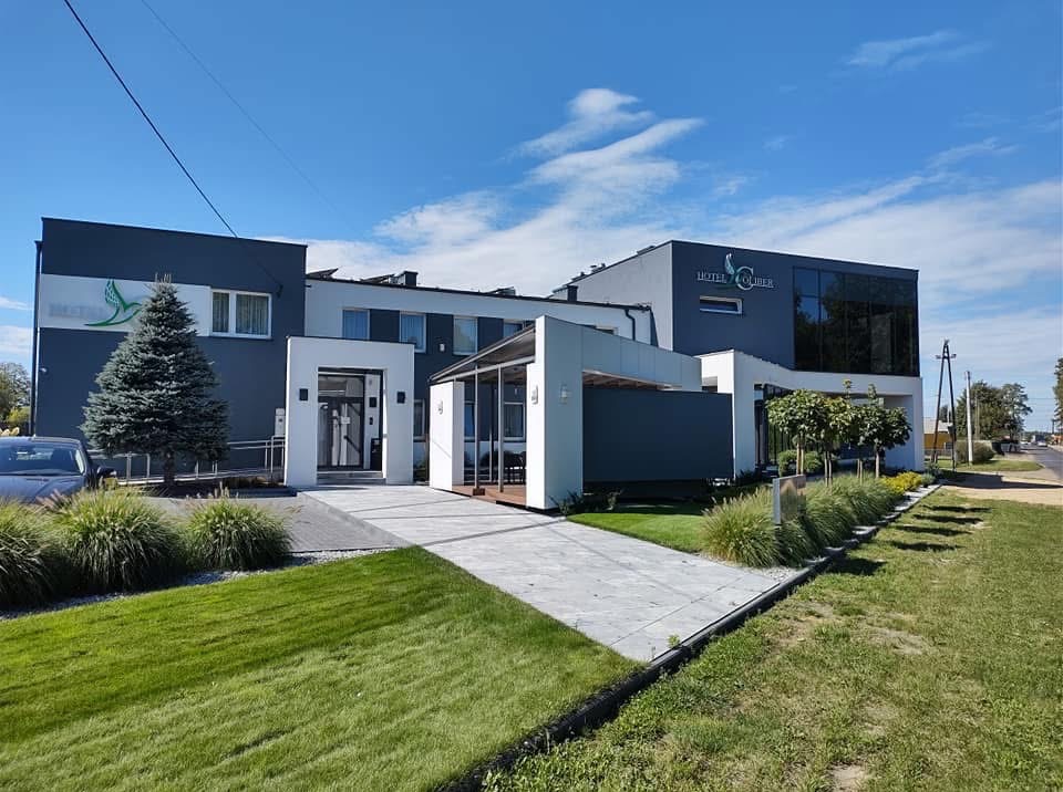 Nowoczesny budynek hotelu z szaro-białą elewacją, widok z zewnątrz w słoneczny dzień, zadbany trawnik i krzewy ozdobne.