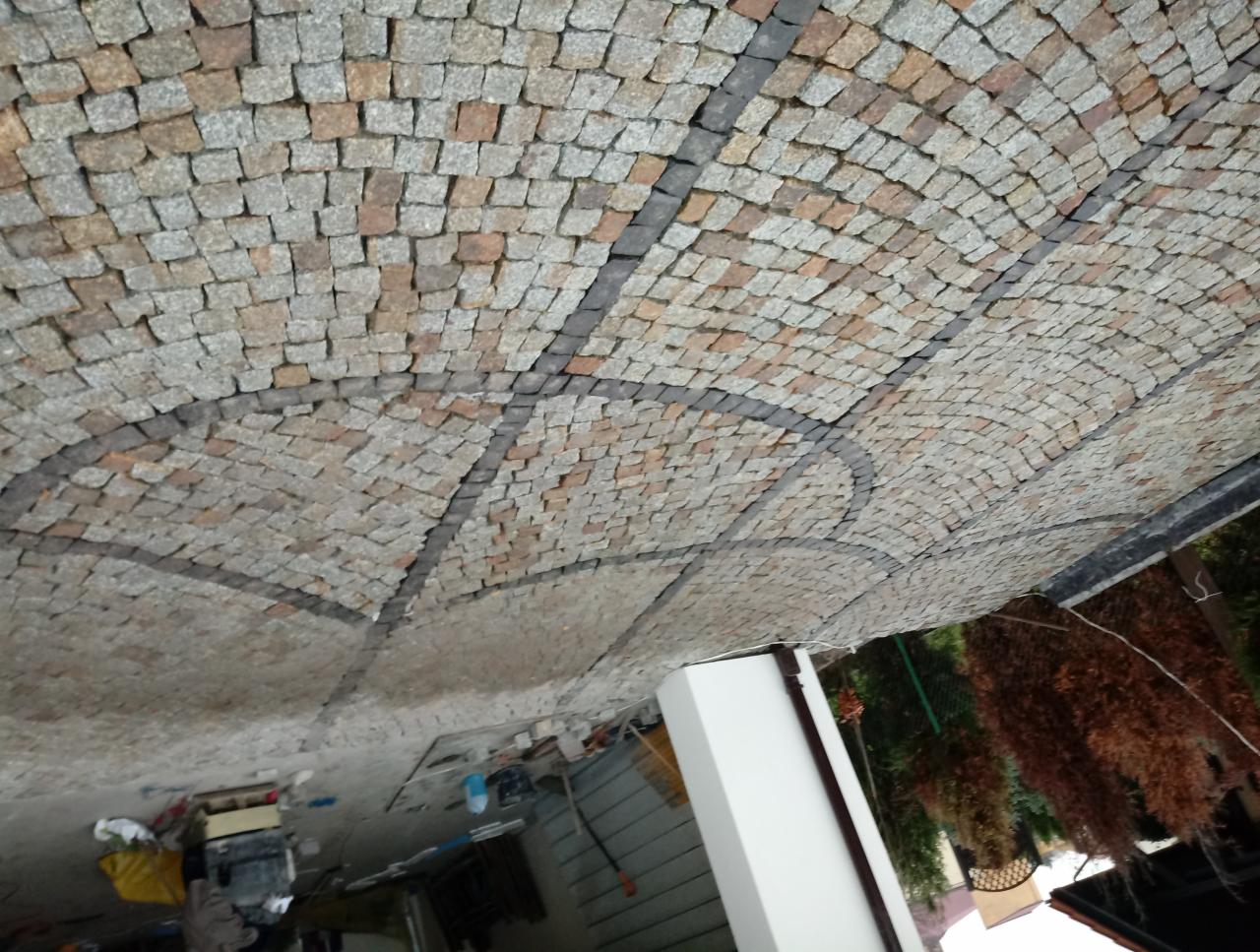 Ułożona kostka granitowa w łukowaty wzór, z ciemniejszymi kostkami tworzącymi dekoracyjne linie. Widoczne niedokończone prace i narzędzia w tle.