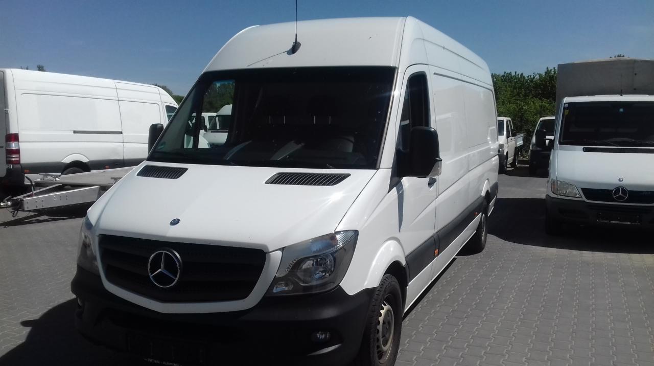 Biały bus marki Mercedes Sprinter zaparkowany na placu, widok z przodu, antena na dachu, w tle inne busy i przyczepa.