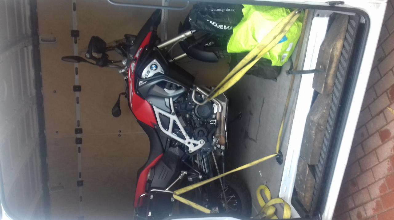 Motocykl BMW GS zabezpieczony pasami transportowymi wewnątrz białego busa, widoczne drewniane kliny stabilizujące.