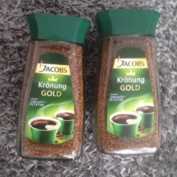 Dwa słoiki kawy rozpuszczalnej Jacobs Krönung Gold z zielonymi wieczkami, ustawione obok siebie na szarym, włochatym dywanie.