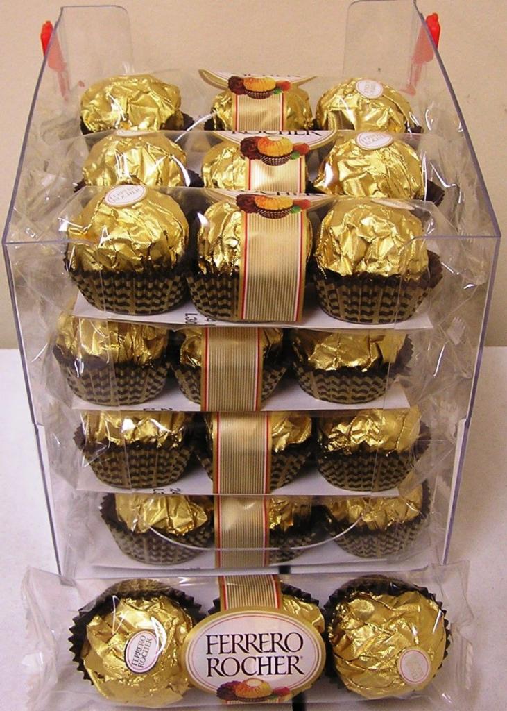 Przezroczysty pojemnik z wieloma czekoladkami Ferrero Rocher w złotych papierkach, ułożonymi w rzędach.