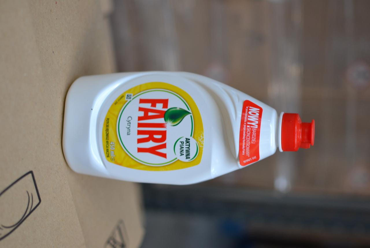 Butelka płynu do mycia naczyń Fairy o zapachu cytrynowym, leżąca na kartonowym podłożu. Widoczna etykieta z logo i informacją o nowej, bardziej skoncentrowanej formule.