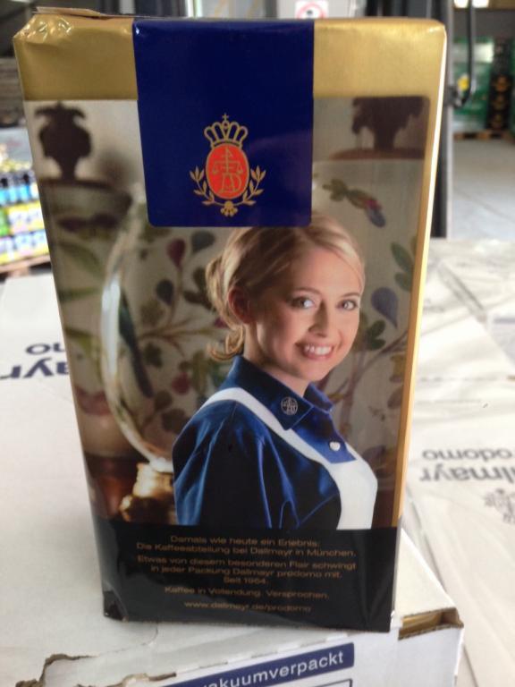 Opakowanie kawy Dallmayr Prodomo z wizerunkiem uśmiechniętej kobiety w mundurze, widoczne logo marki i tekst informacyjny w języku niemieckim, ustawione na kartonie w magazynie.
