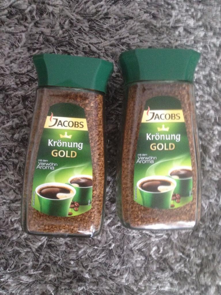 Dwa słoiki kawy rozpuszczalnej Jacobs Krönung Gold z zielonymi wieczkami, ustawione obok siebie na szarym, włochatym dywanie.