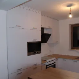 Nowoczesna kuchnia z białymi szafkami z minimalistycznymi uchwytami, wbudowanym piekarnikiem, czarnym okapem i drewnianym blatem, oświetlona wiszącą żarówką.