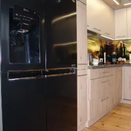Nowoczesna kuchnia z czarną lodówką side-by-side z dystrybutorem wody i frontami szafek w odcieniach drewna, oświetlona listwami LED.