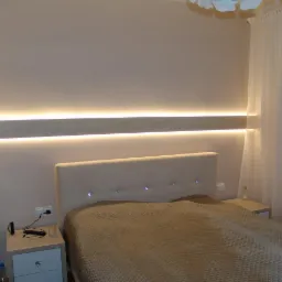 Nowoczesna sypialnia z beżowym łóżkiem, podświetlanym panelem ściennym i zwiewnymi białymi zasłonami.