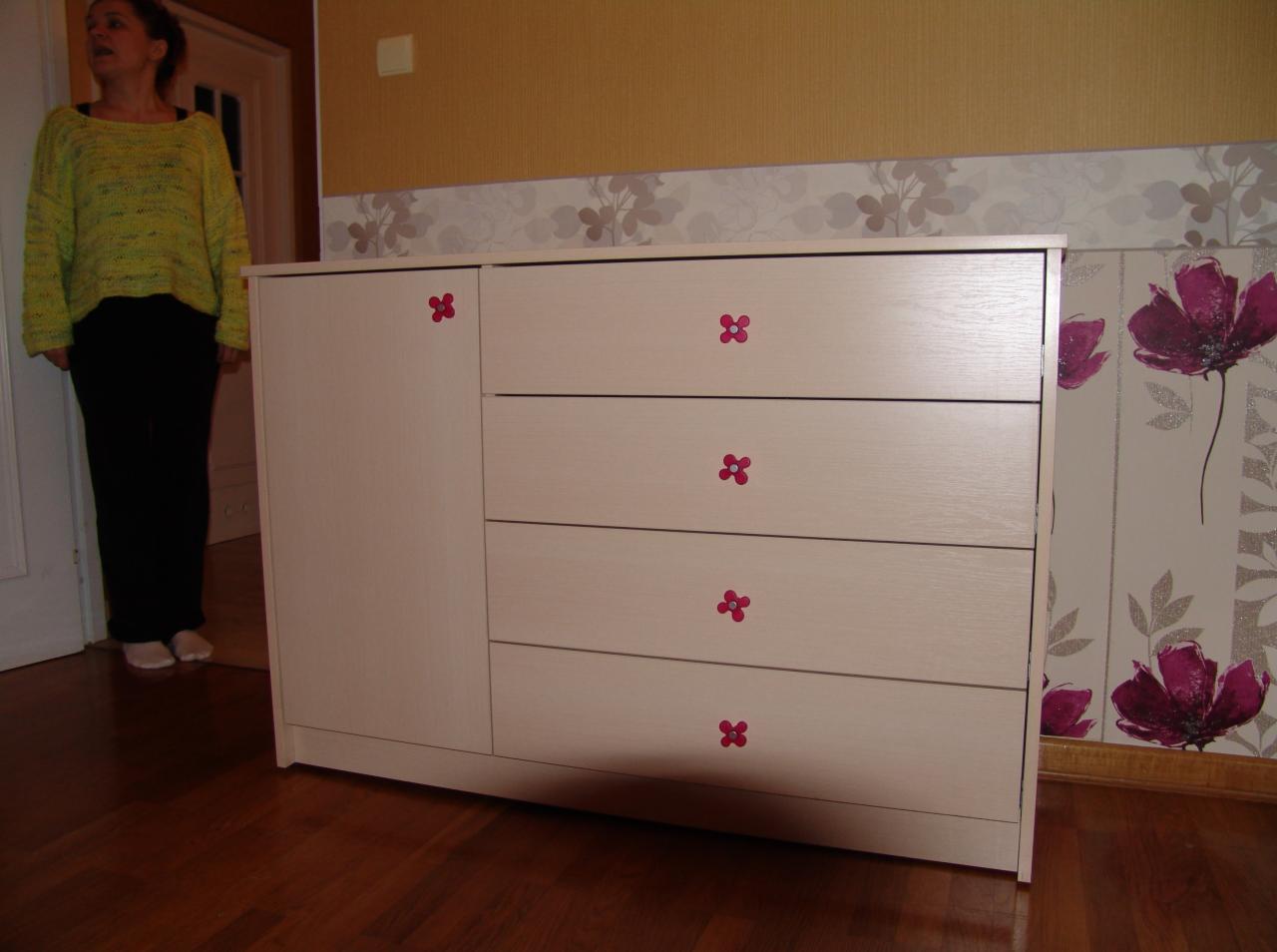 Jasna komoda z czterema szufladami i szafką boczną, ozdobiona różowymi kwiatkami, na tle wzorzystej tapety i drewnianej podłogi, z widoczną częściowo kobietą w tle.