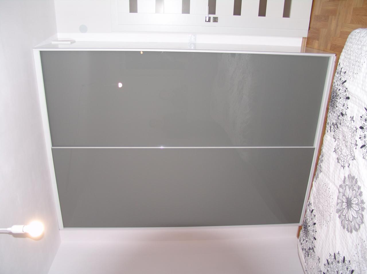Szara szafa przesuwna z białymi obramowaniami, widoczna z dołu, obok łóżka z wzorzystą pościelą i białych drzwi.