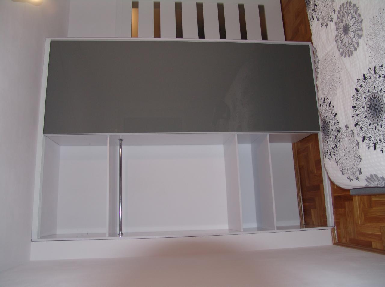 Wbudowana szafa z szarymi przesuwnymi drzwiami i białymi półkami w sypialni, widok z góry, z boku widoczny fragment łóżka z wzorzystą pościelą i drewniana podłoga.