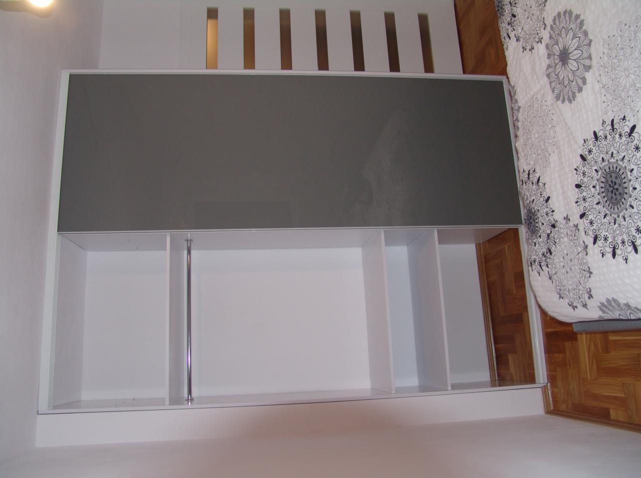 Wnętrze szafy wnękowej z szarymi przesuwnymi drzwiami, widoczne półki i metalowy drążek, obok łóżko z wzorzystą pościelą i parkiet.