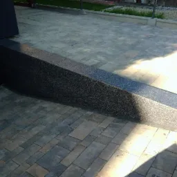 Ułożona kostka brukowa w odcieniach szarości, z ciemnoszarą, wykończoną mozaiką rampą prowadzącą do ściany. Widoczne cienie i fragment metalowego ogrodzenia w tle.