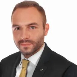 Portret młodego mężczyzny z lekkim zarostem, ubranego w ciemny garnitur i złotą krawat, na białym tle.