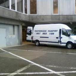 Biały bus z otwartymi drzwiami zaparkowany na parkingu przed budynkiem z numerem telefonu i napisem 'Przeprowadzki Transport'.