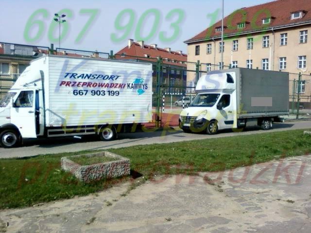 Dwa samochody dostawcze na tle budynku mieszkalnego i boiska szkolnego. Na jednym z aut widnieje napis 'Transport Przeprowadzki Kammys'.
