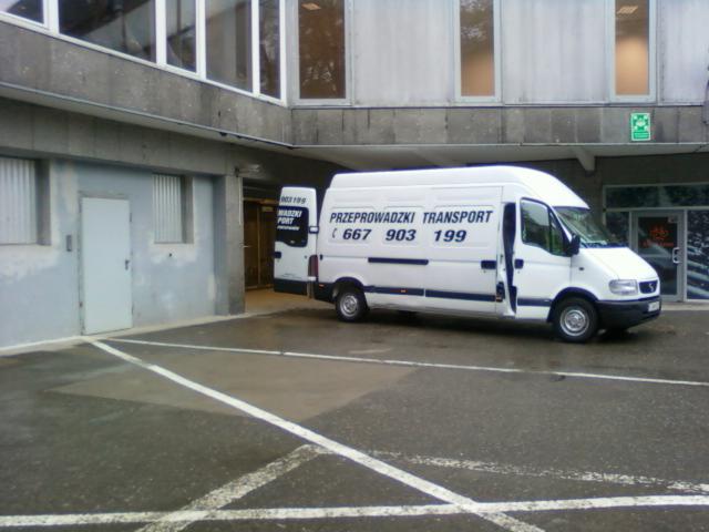 Biały bus z otwartymi drzwiami zaparkowany na parkingu przed budynkiem z numerem telefonu i napisem 'Przeprowadzki Transport'.