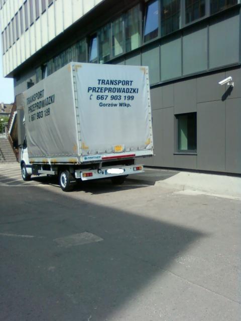 Samochód dostawczy z plandeką z napisem 'Transport Przeprowadzki 667 903 199 Gorzów Wlkp.' zaparkowany przed nowoczesnym budynkiem biurowym.