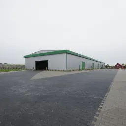 Szeroki, parterowy magazyn z szarą elewacją i zielonym dachem, widoczny z zewnątrz, z otwartą bramą wjazdową i utwardzonym placem przed budynkiem.