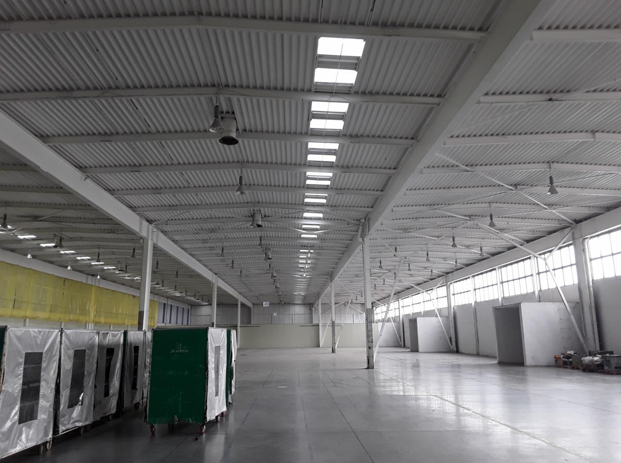 Wnętrze przestronnego magazynu z białym, falistym dachem, oświetlonym przez świetliki i lampy, z widocznymi wózkami do transportu towarów po lewej stronie i oknami w tle.