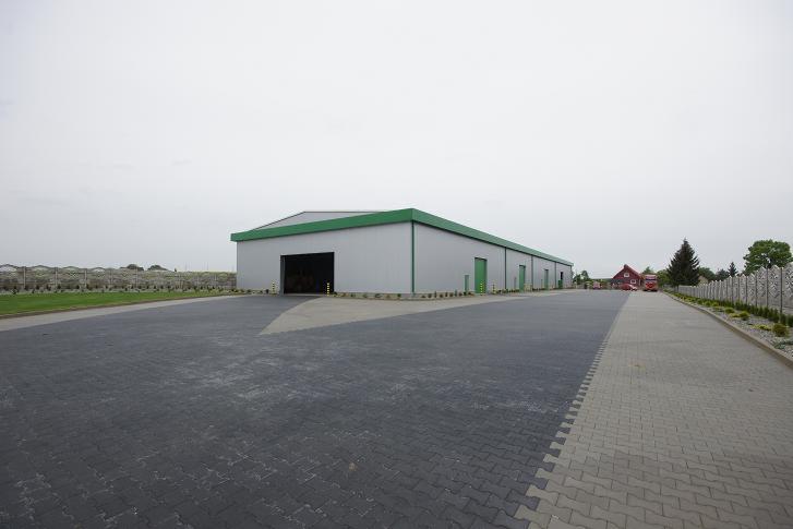 Szeroki, parterowy magazyn z szarą elewacją i zielonym dachem, widoczny z zewnątrz, z otwartą bramą wjazdową i utwardzonym placem przed budynkiem.