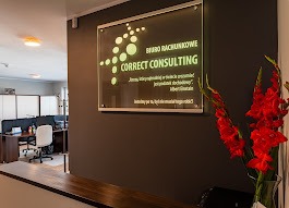 Tablica informacyjna z nazwą firmy 'Correct Consulting' i cytatem Alberta Einsteina, umieszczona na ścianie w biurze, obok wazon z czerwonymi mieczykami i widok na stanowiska pracy.