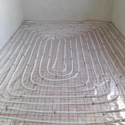 Białe rury ogrzewania podłogowego ułożone spiralnie na folii z czerwoną siatką w surowym pomieszczeniu z widocznym oknem.