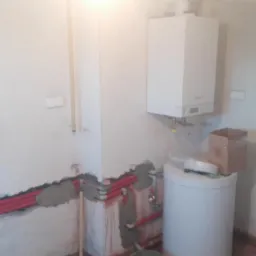 Wnętrze w trakcie remontu z widocznymi czerwonymi rurami instalacji grzewczej w ścianie, białym kotłem gazowym na ścianie i białym zasobnikiem wody na podłodze, oraz kartonowym pudełkiem obok.