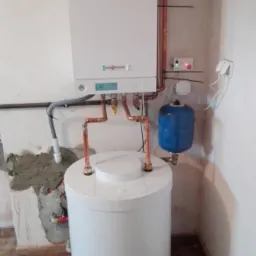 Nowoczesny, biały piec gazowy kondensacyjny z zasobnikiem ciepłej wody, zamontowany na tle surowej ściany z widocznymi śladami po kuciu i drewnopodobną podłogą.