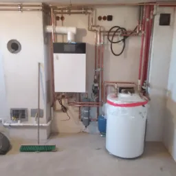 Nowoczesny piec gazowy wiszący, podłączony do instalacji z miedzianych rur, obok biały zasobnik ciepłej wody zabezpieczony folią, widoczny również filtr, grzejnik i szczotka do sprzątania.