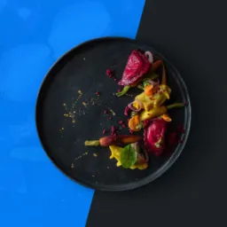 Elegancka kompozycja pierożków w różnych kolorach (czerwone, żółte, pomarańczowe) z warzywami i ziołami na czarnym talerzu, na tle podzielonym na niebieski i czarny obszar.