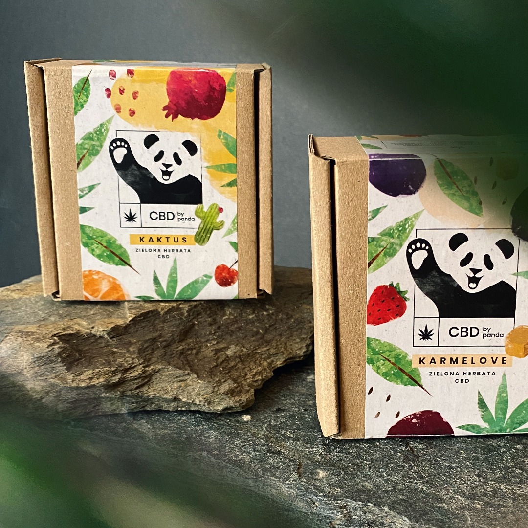 Dwa kartonowe pudełka z etykietami 'CBD by panda' na kamiennej powierzchni, etykiety z motywami owoców, roślin i pandy z podniesioną łapą, jeden o smaku 'Kaktus' drugi 'Karmelove'.