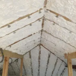 Poddasze z drewnianą konstrukcją pokryte białą pianą izolacyjną, widoczne metalowe uchwyty mocujące, fragment okna w tle.