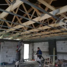 Montaż metalowego rusztu pod płyty gipsowo-kartonowe na drewnianej konstrukcji dachu, widoczni dwaj pracownicy w trakcie pracy, z narzędziami i drabiną w surowym wnętrzu budynku z białych pustaków.